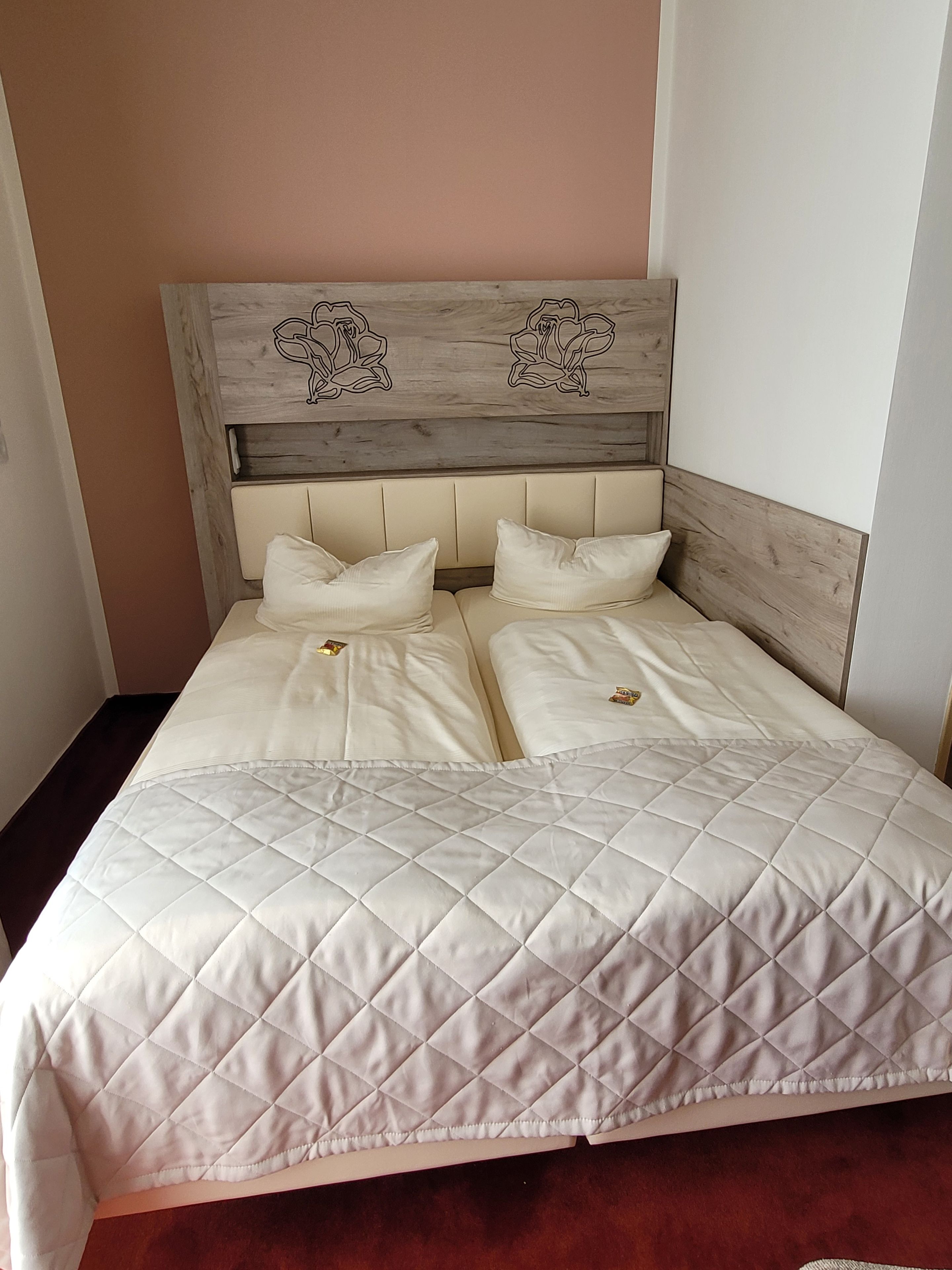 Un lit double avec une couette blanche matelassée et deux oreillers, une tête de lit en bois ornée de motifs floraux sculptés, et un mur sobre en arrière-plan.