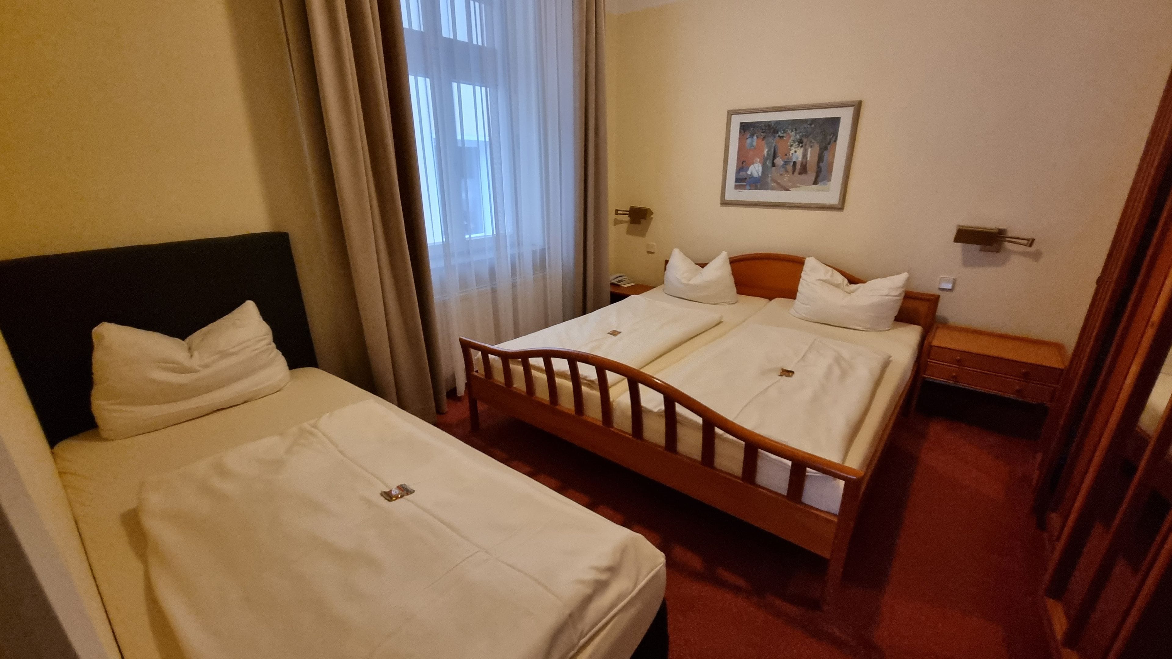 Habitación de hotel con dos camas individuales, cabecera de madera, un cuadro en la pared y una mesita de noche con lámpara.