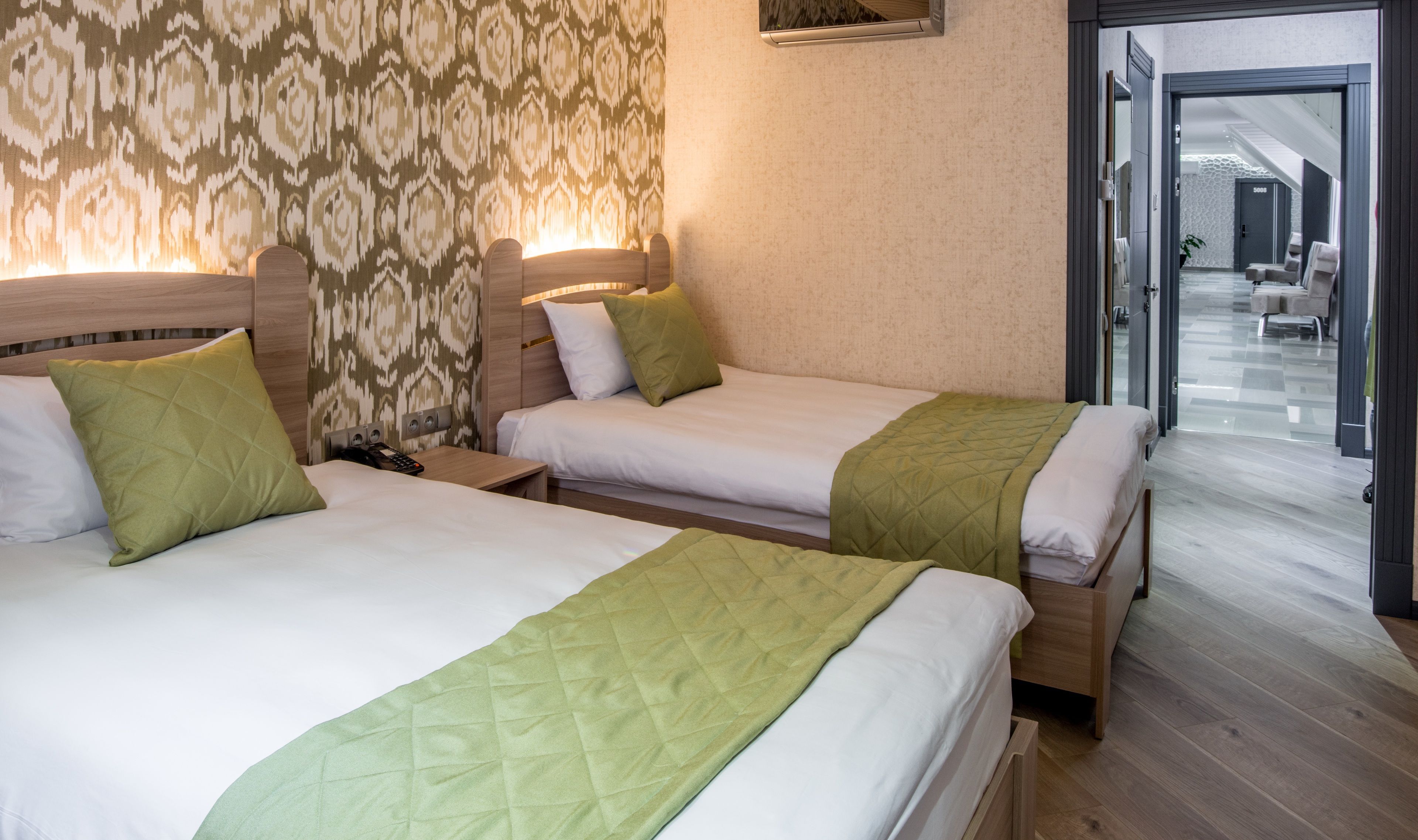 A hotel room with two beds, each with green bedspreads and white linens. There is a patterned wall behind the beds and a door leading to a hallway with seating areas.