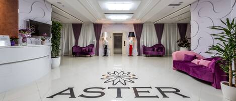 Un vestíbulo de hotel con una recepción, sofás morados y un letrero que dice "ASTER HOTEL GROUP".