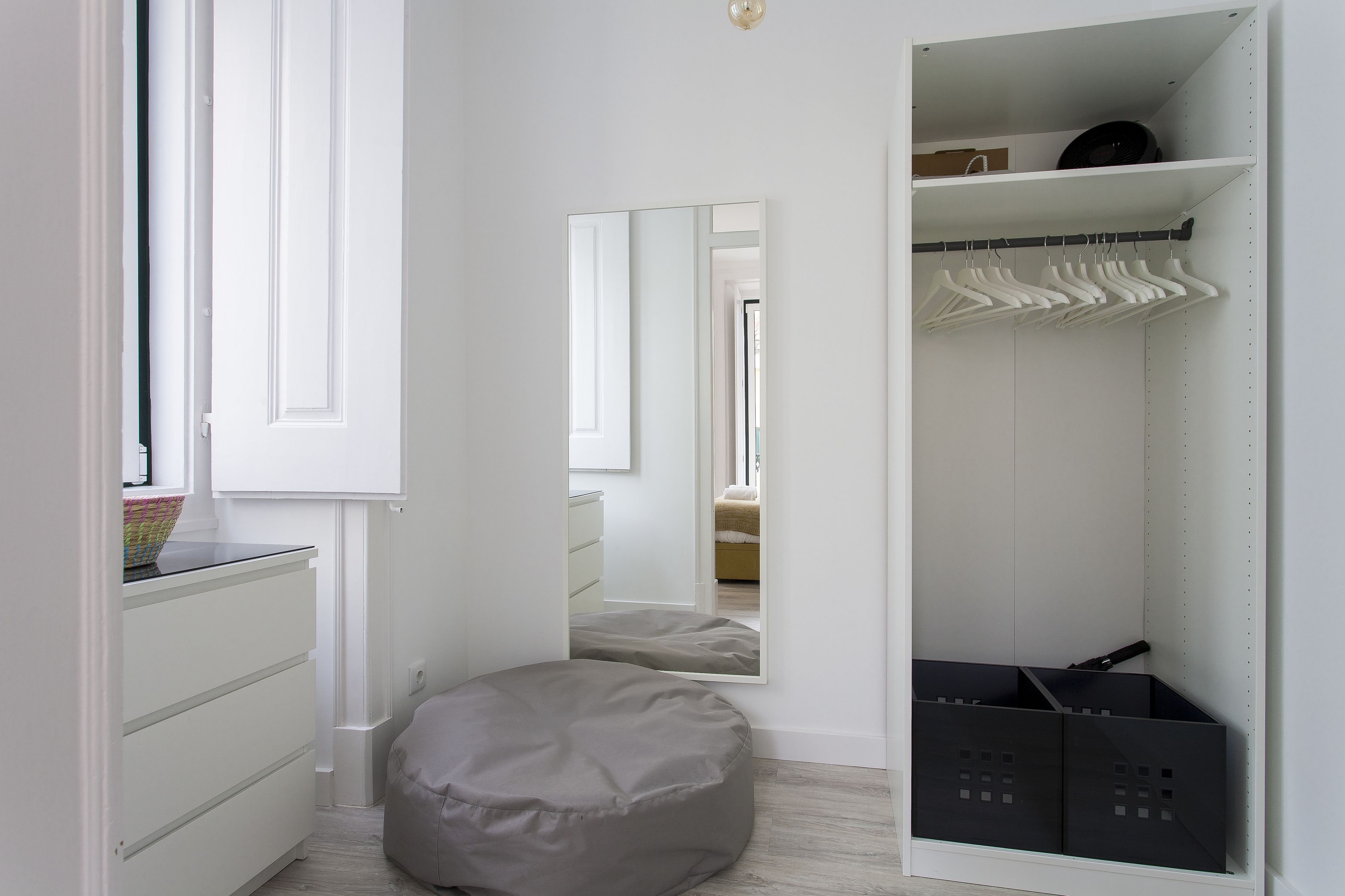 Een slaapkamer met een witte linnenkast, een ronde relaxfauteuil, een spiegel en een inloopkast met hangende kleding en opbergdozen.