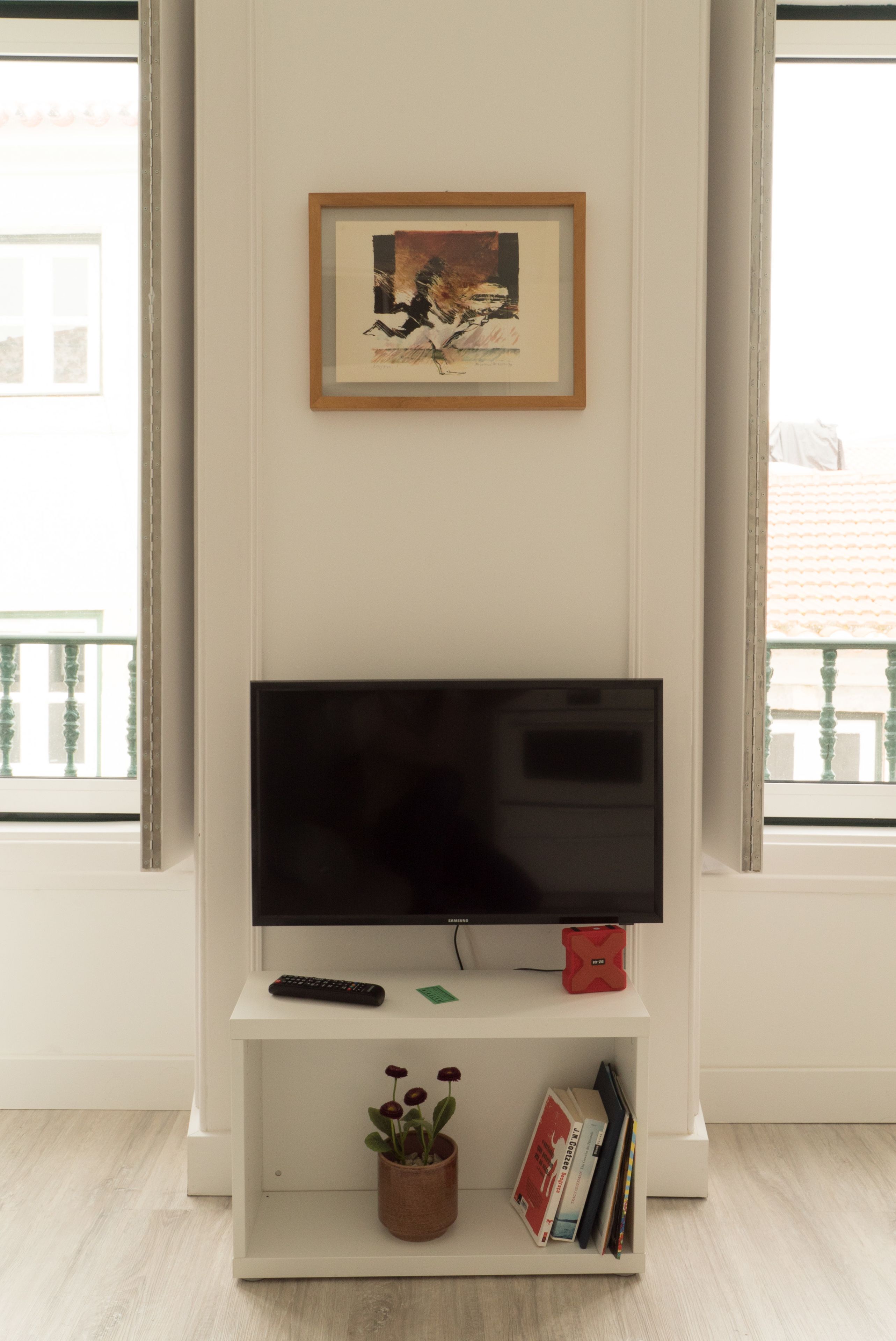 Una sala de estar con un televisor de pantalla plana sobre un soporte blanco, un cuadro en la pared, una planta en maceta y libros sobre el soporte.