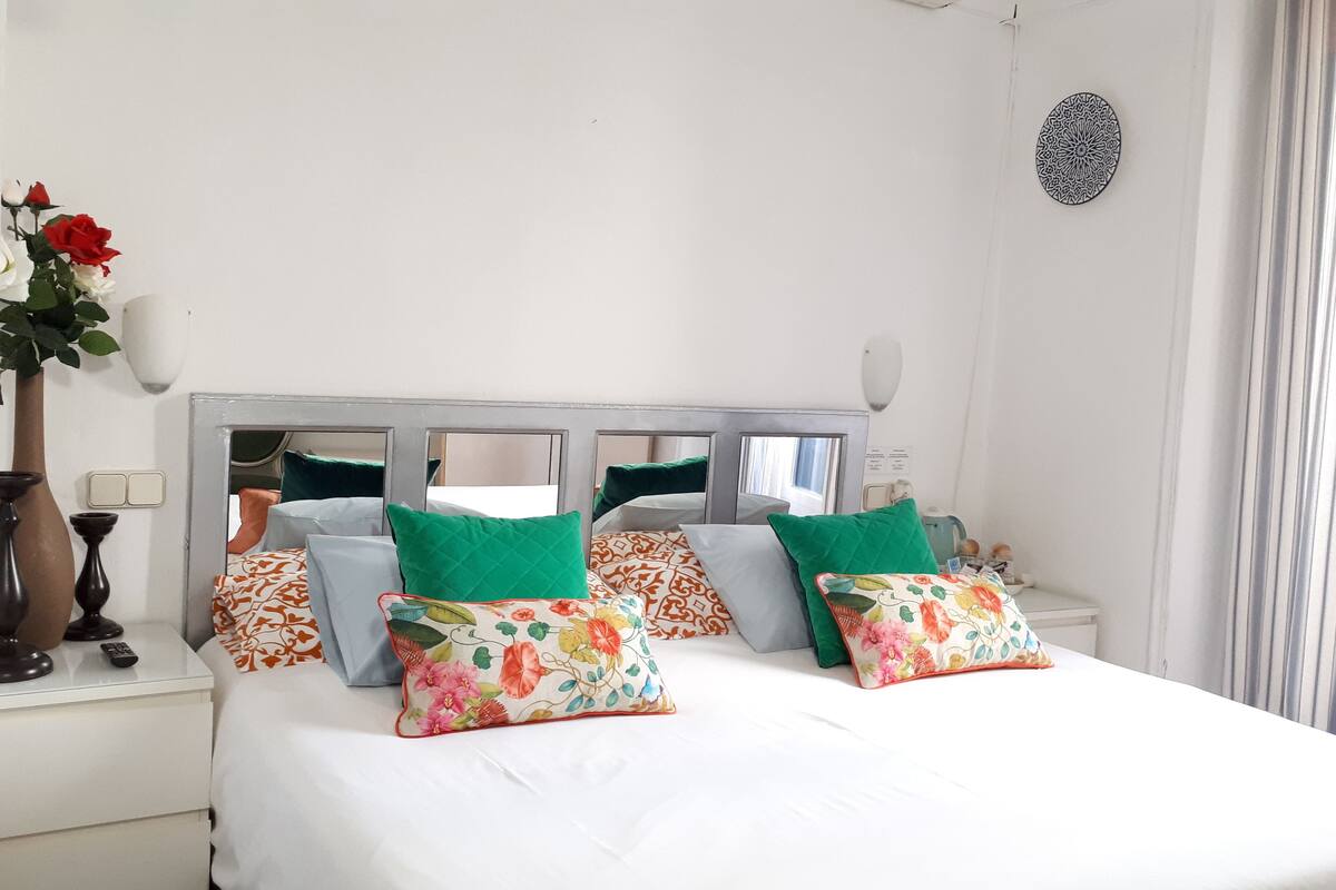 A neatly arranged bedroom with a large bed, multiple pillows, a bedside table with a vase of flowers, and a mirror on the headboard.