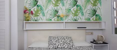Une chambre à coucher avec un lit, une table de chevet, une étagère avec un tableau, et un mur orné d’un motif de feuilles.