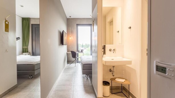Habitación de hotel moderna con cama, escritorio, silla, baño con lavamanos y espejo, y vistas al exterior.