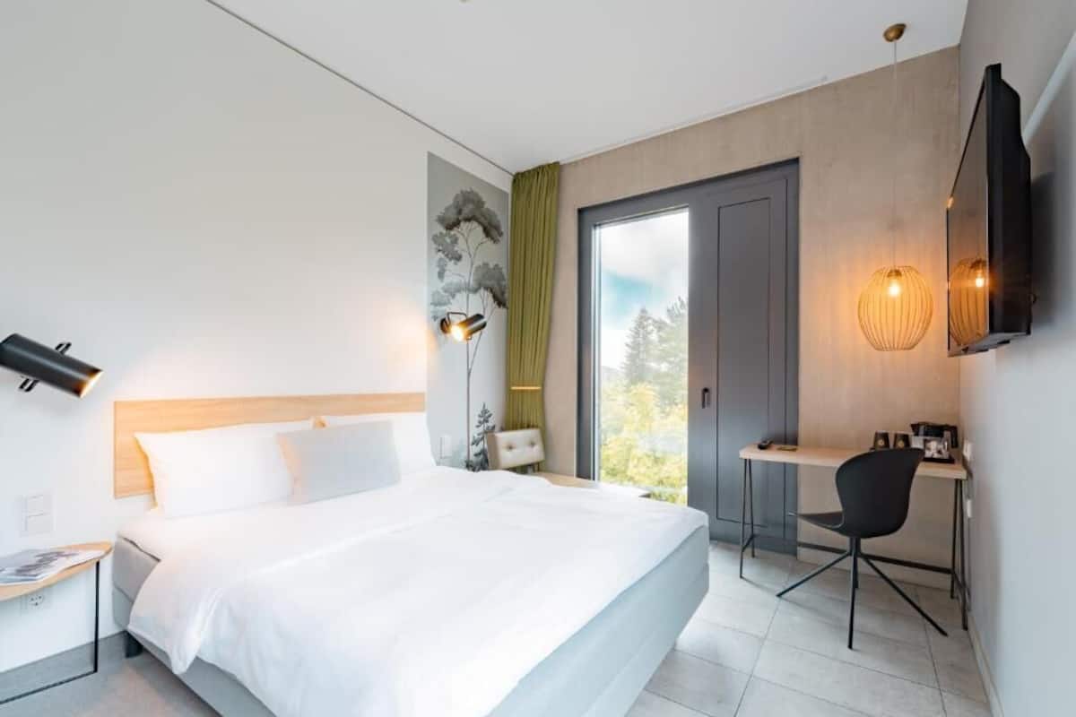 Een moderne hotelkamer met een groot bed, een bureau, een stoel, een televisie en een raam met uitzicht op bomen.