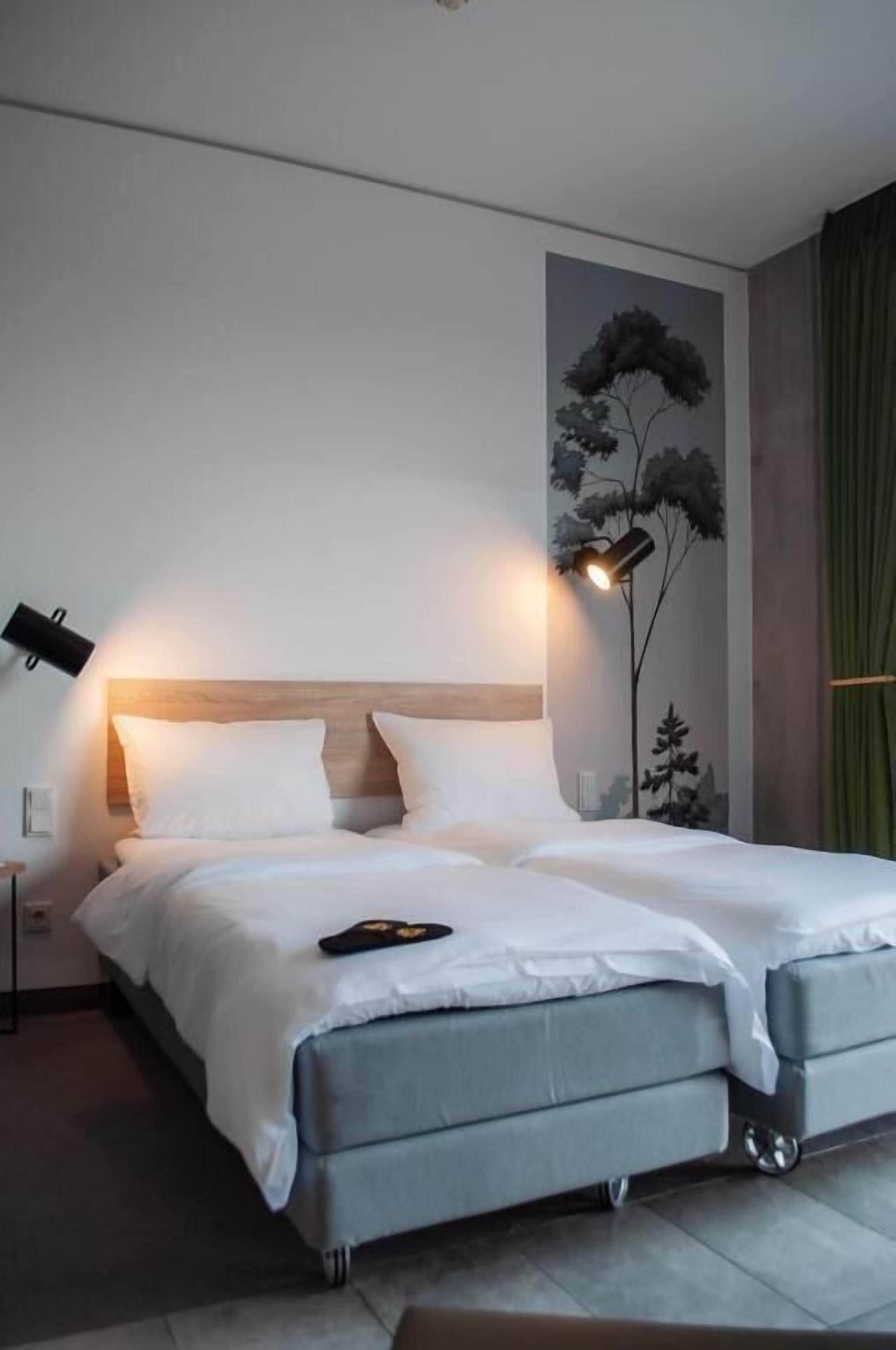 Un lit double avec une tête de lit en bois, deux lampes de chevet, un téléviseur fixé au mur et une fresque représentant des arbres.