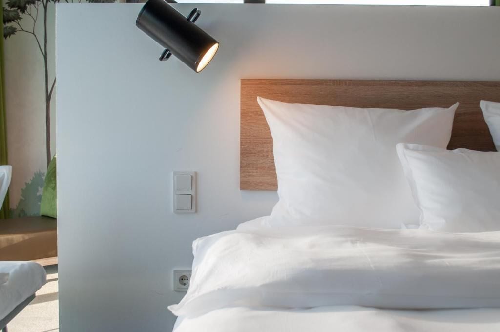 A neatly made bed with white linens, a wooden headboard, and a wall-mounted light fixture.