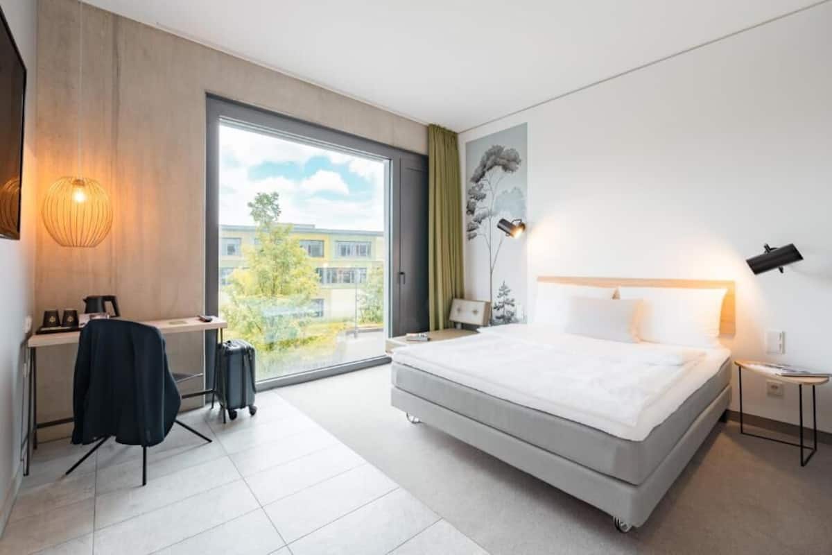 Een moderne hotelkamer met een groot bed, een bureau, een stoel, een koffer, een lamp en uitzicht op gebouwen.