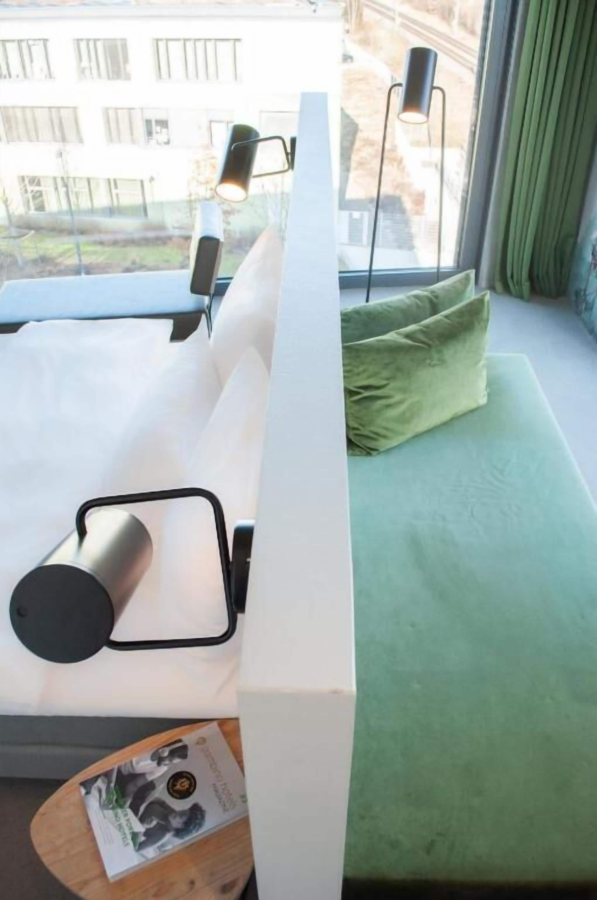 Ein modernes Zimmer mit einem weißen Bett, grünen Kissen, einem Nachttisch mit einer Zeitschrift und Blick auf Gebäude durch ein Fenster.