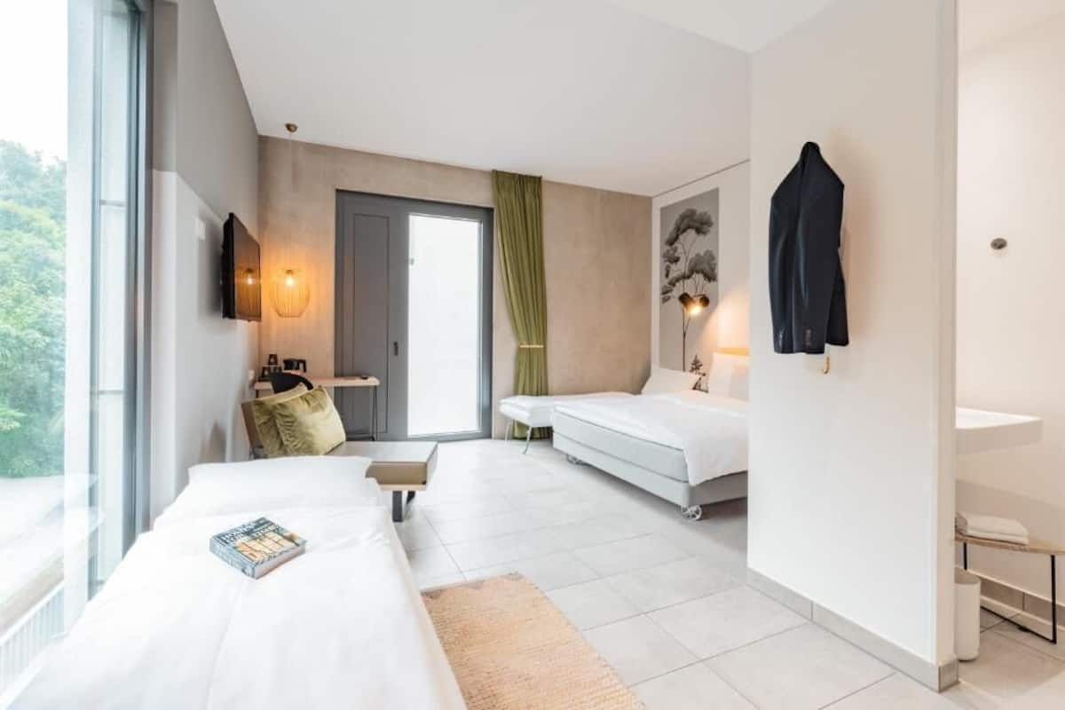 Een moderne hotelkamer met een groot raam, een bed, een nachtkastje, een bureau, een stoel, een bank, een televisie, een lamp en een kapstok.