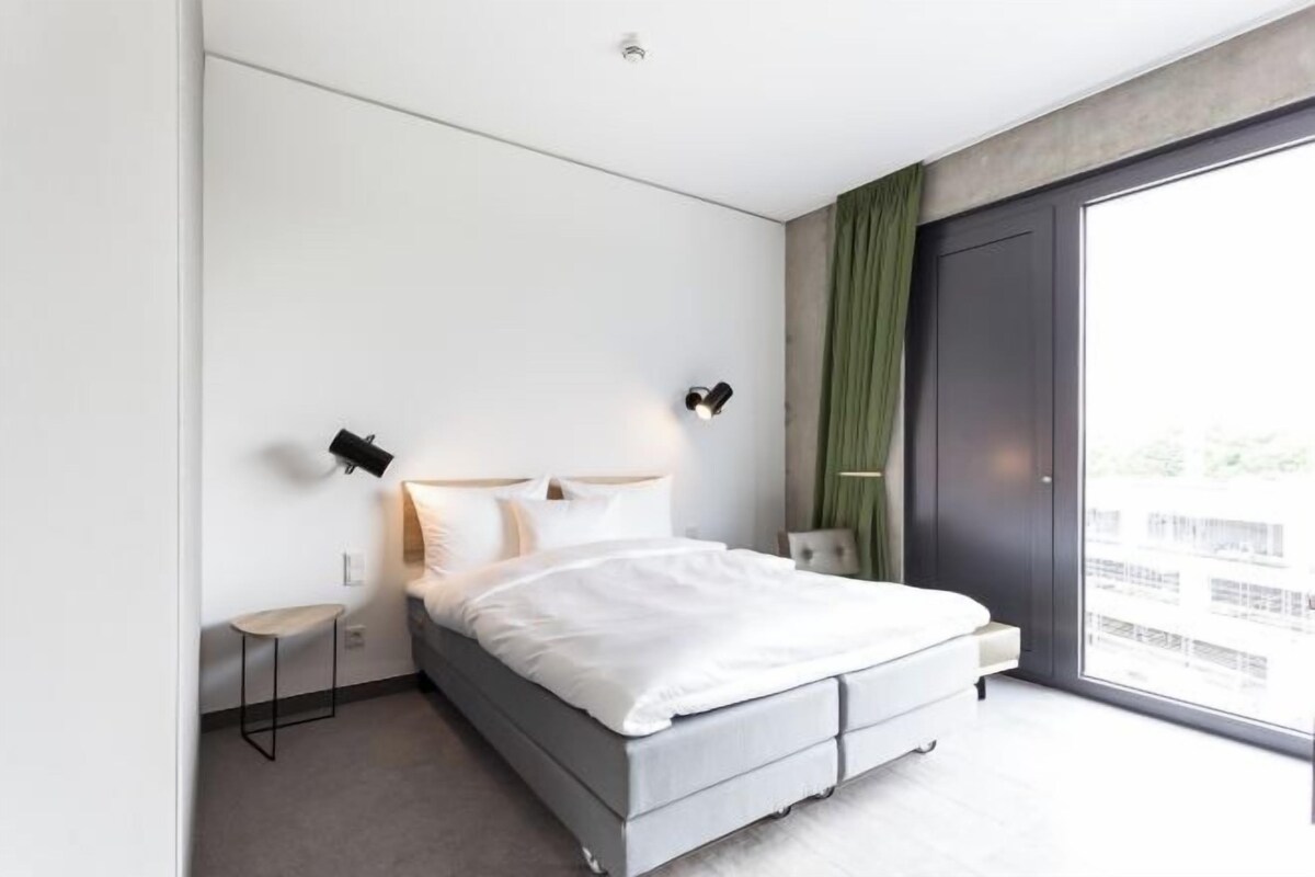Una habitación de hotel moderna con una cama grande, una mesita de noche pequeña, una lámpara de pared y vistas amplias a través de un ventanal.