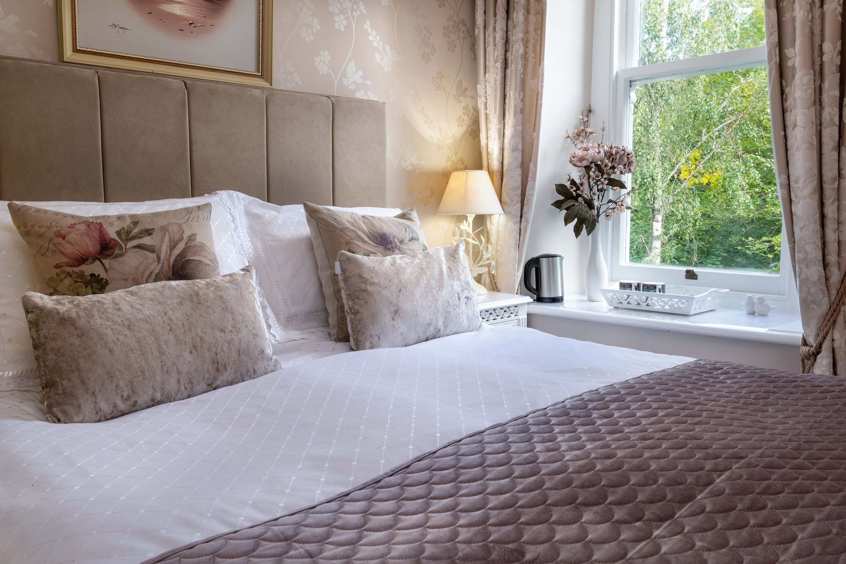 A neatly made bed with a quilted headboard, a bedside table with a lamp, and a window with curtains.