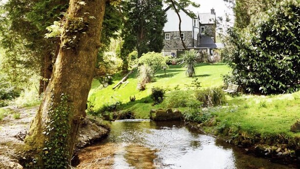 A house with a stone facade, surrounded by lush greenery and a small stream.