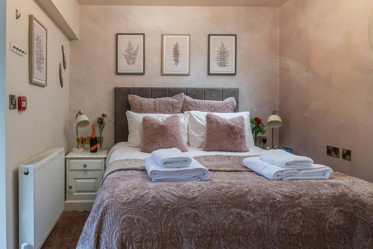 A neatly made bed with a patterned bedspread and pillows, a bedside table with a lamp and a bottle, and framed botanical prints on the wall.