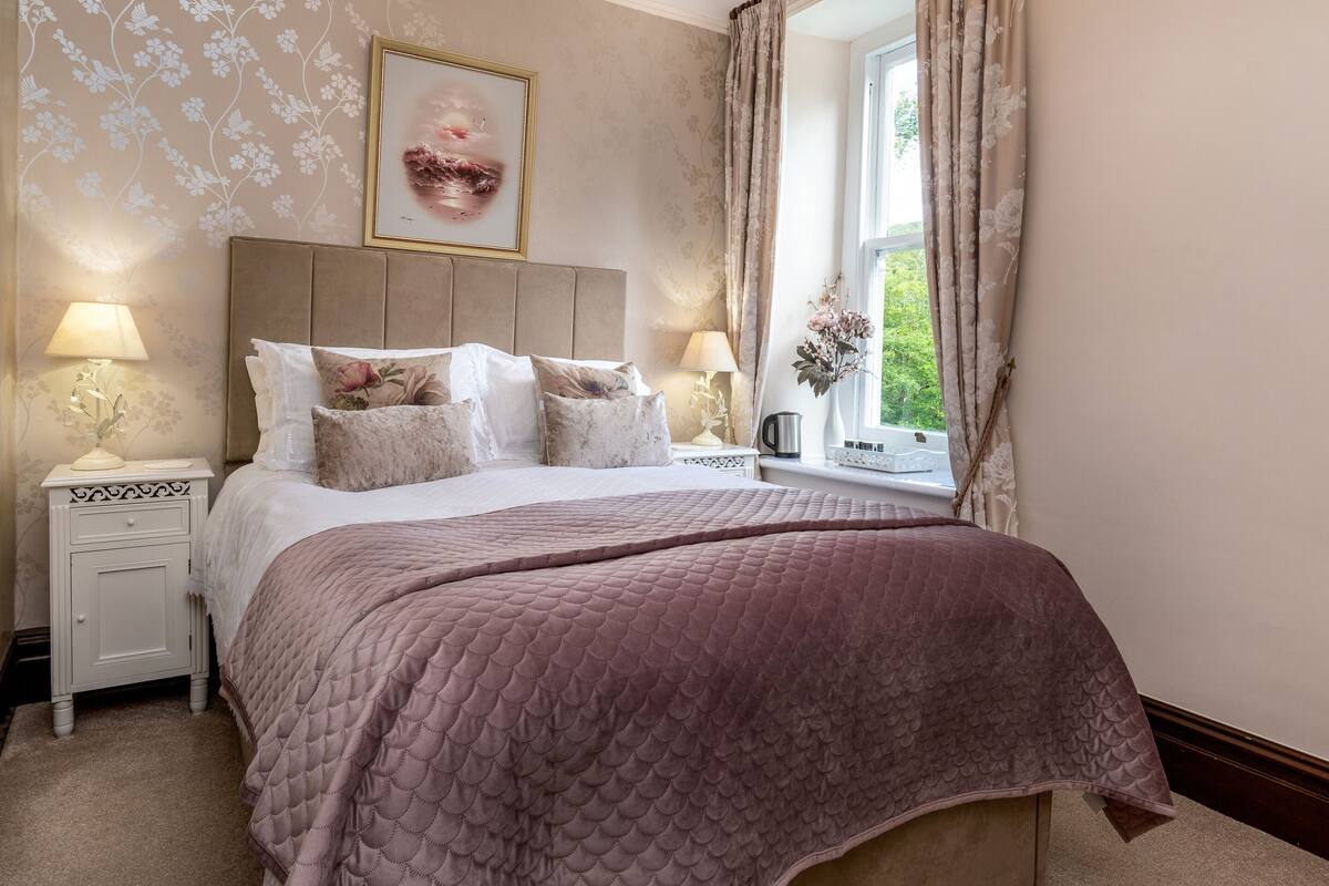 A bedroom with a large bed, two bedside tables with lamps, a window with curtains, and a floral wall design.