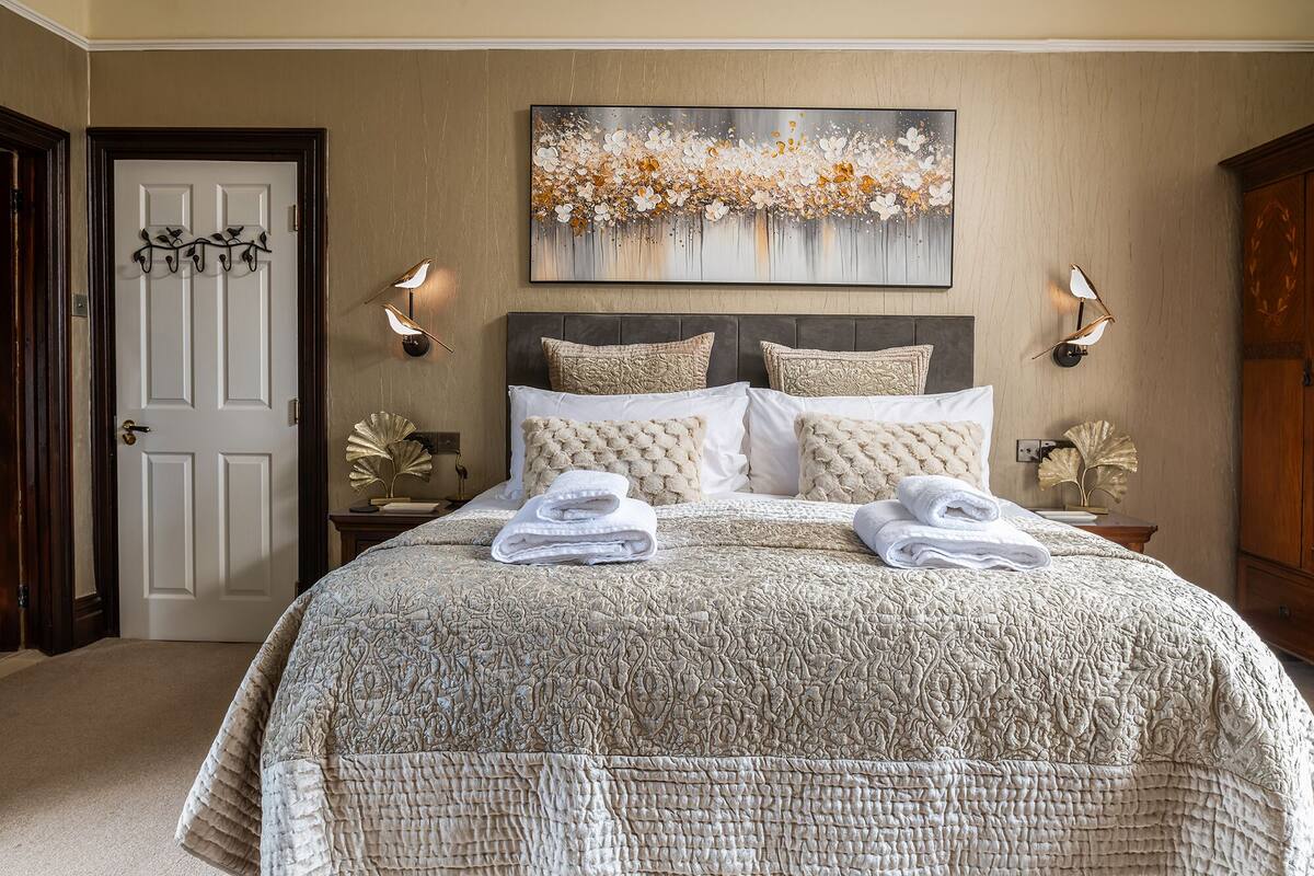 A bedroom with a large bed, two wall-mounted lamps, a painting above the bed, and a door with a coat rack.