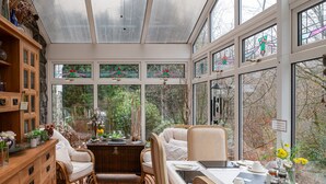 A conservatory with a wooden cabinet, wicker chairs, a dining table with tea set, and a view of trees through large windows.