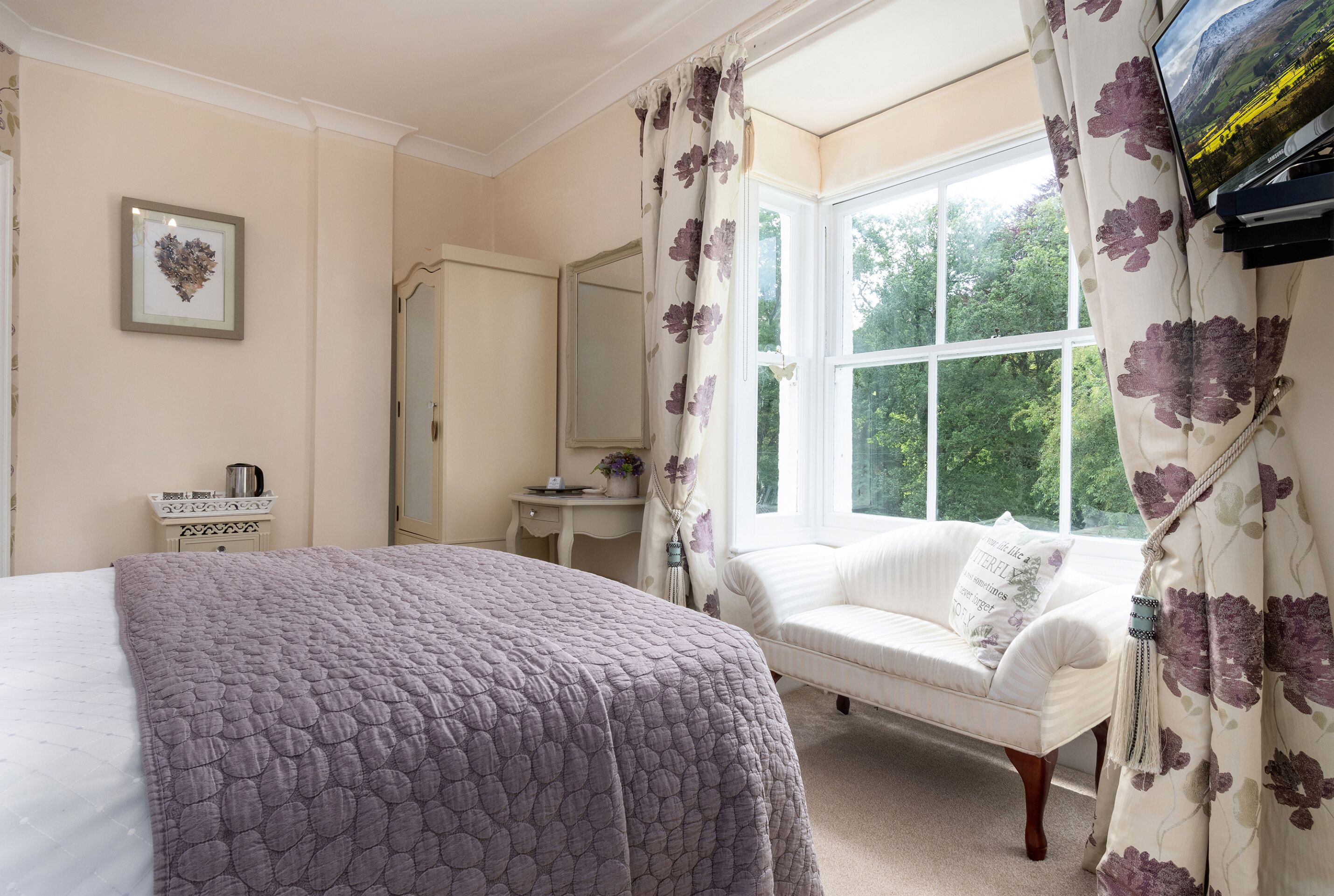 A bedroom with a large bed, a white sofa, a window with curtains, a mirror, and a television on a stand.