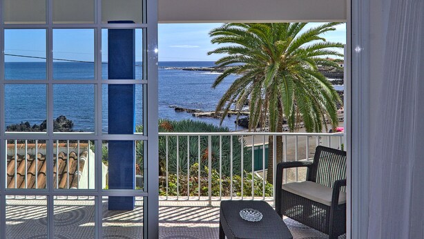 Un balcón con vista al mar, una palmera y una silla de mimbre.