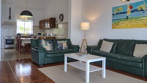 Un salón espacioso con un sofá verde, una mesa de centro blanca y una zona de comedor con una cocina al fondo.