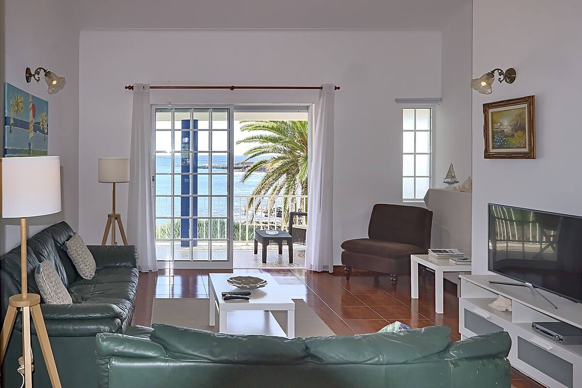 A living room with a green sofa, a brown armchair, a coffee table, a TV, and a balcony with a view of the sea.