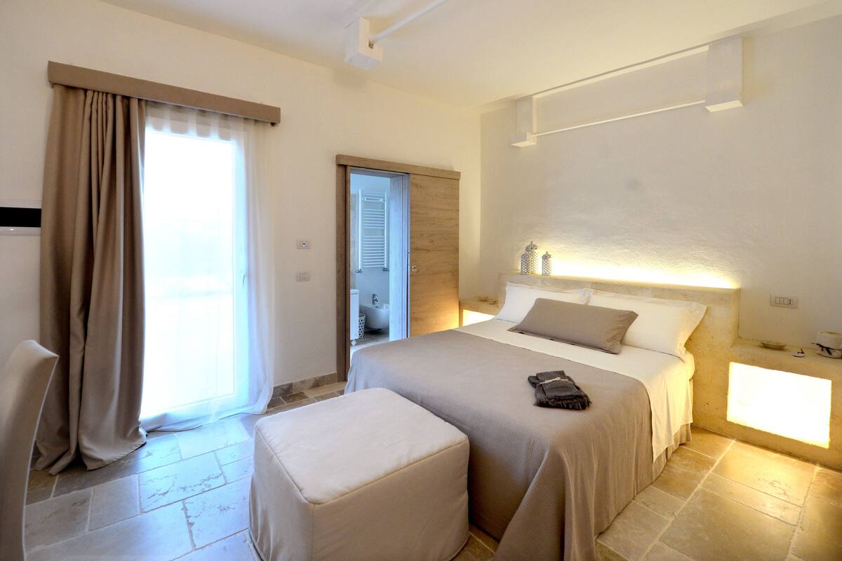 Una habitación de hotel con cama, una silla, una mesita y un ventanal con cortinas.