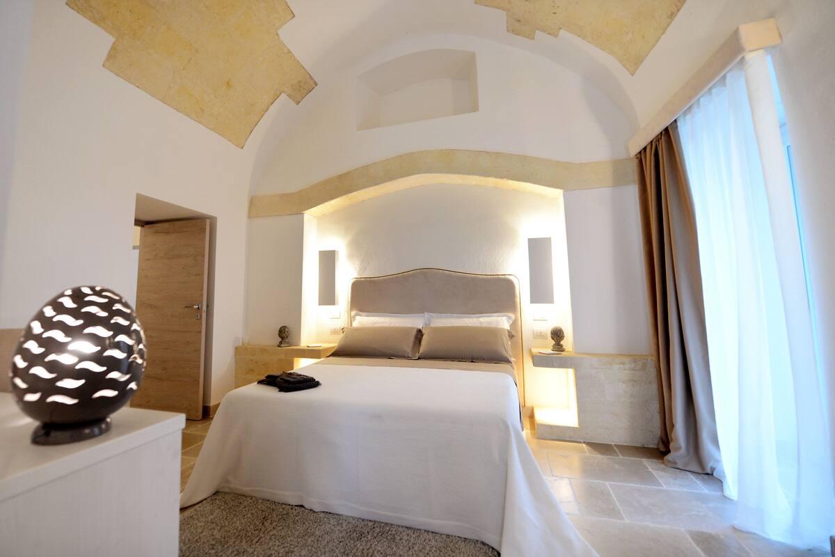 Habitación de hotel con una cama grande, un objeto esférico decorativo en una mesita de noche y vistas al exterior a través de una ventana con cortinas.