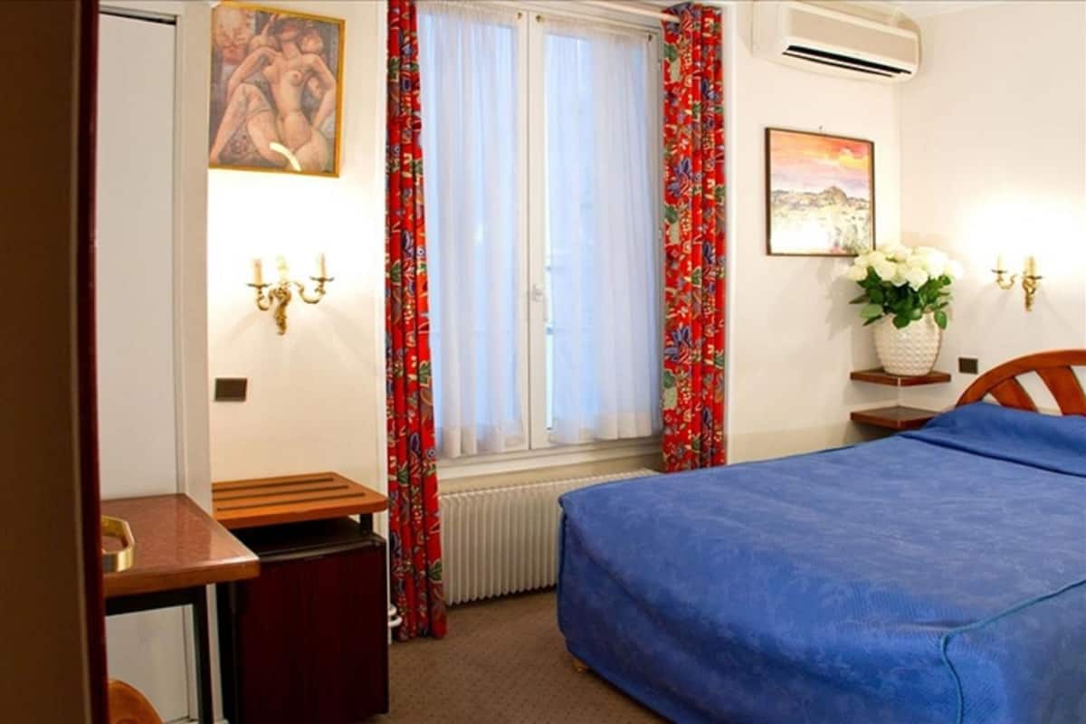 Una habitación de hotel con cama, escritorio, silla, radiador, ventana con cortinas y aire acondicionado instalado en la pared.