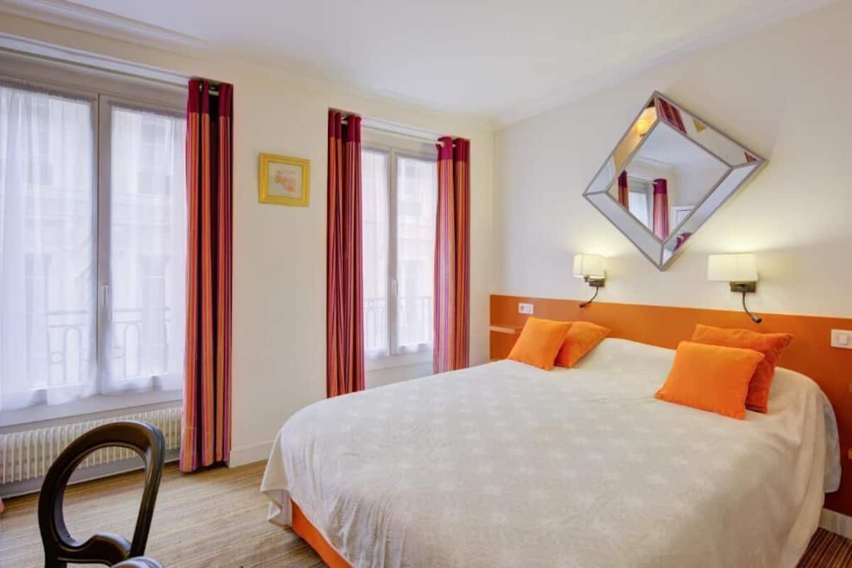 Una habitación de hotel con una cama grande, dos lámparas de noche, un espejo y un ventanal con cortinas.