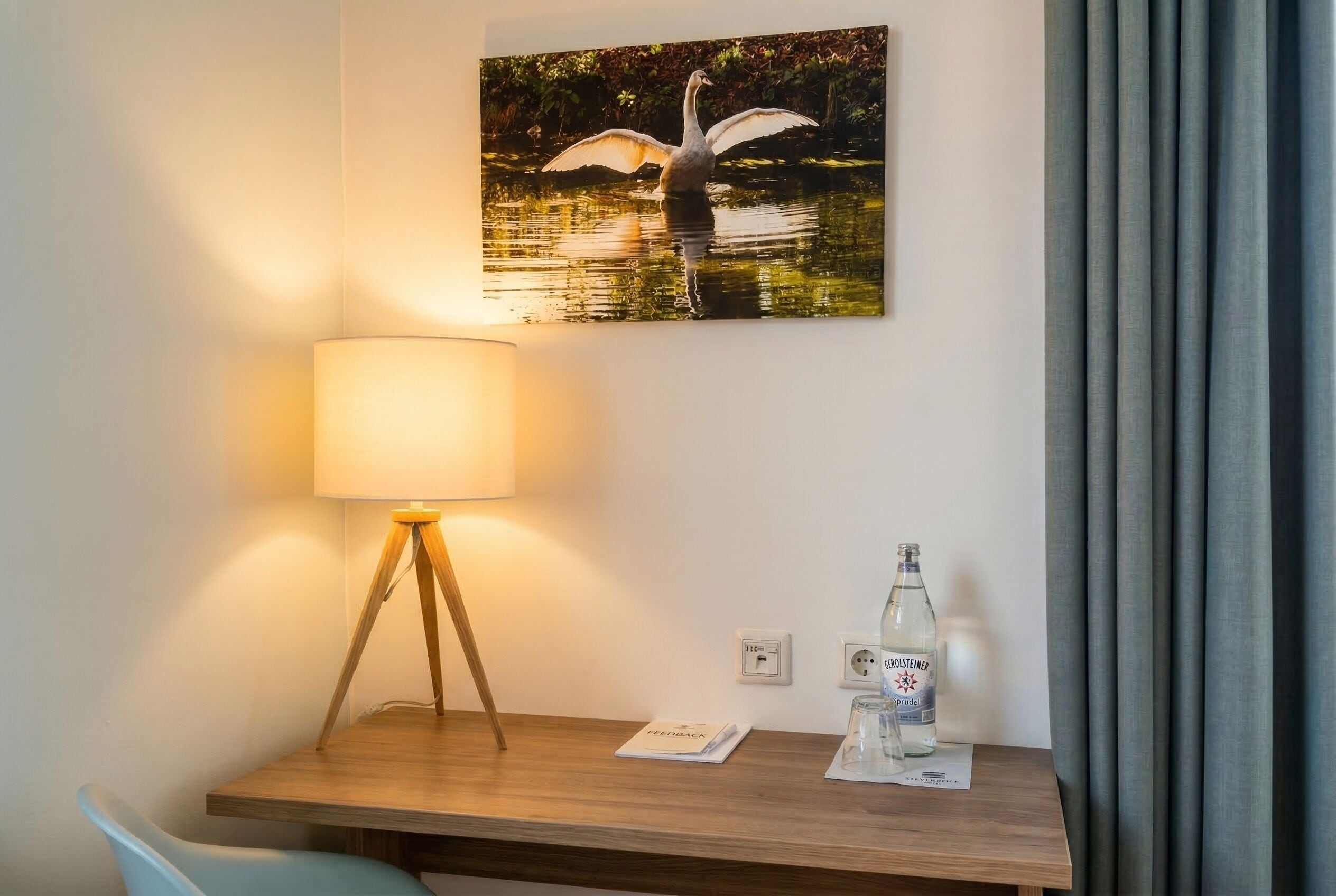 A hotel room with a wooden desk, a lamp, a bottle of water, and a framed picture of a swan on the wall.