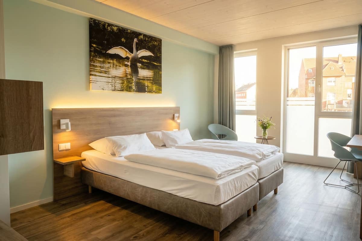 A double bed with a wooden headboard, a nightstand, a wall-mounted lamp, a chair, and a window with curtains.