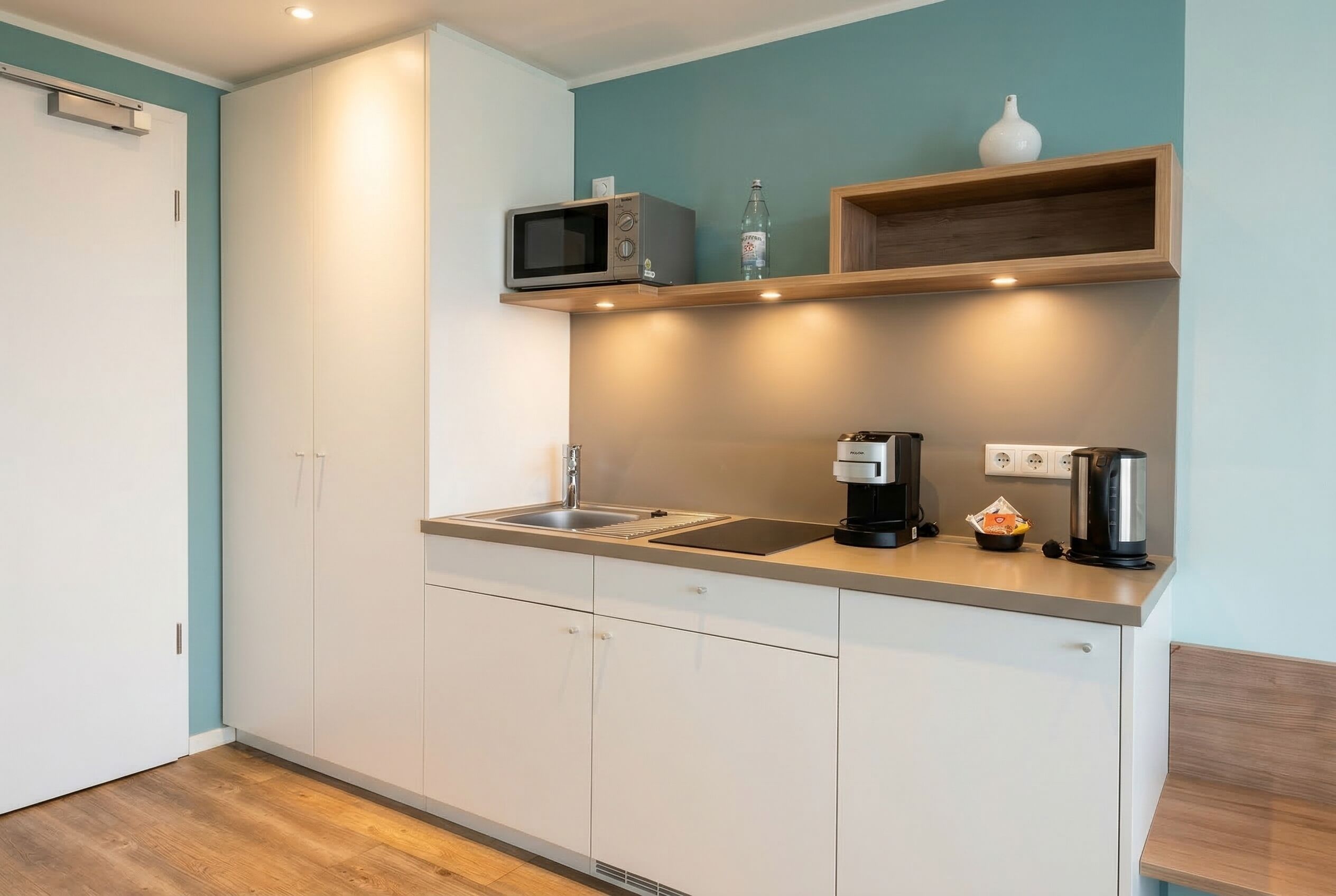 Una cocina moderna con armarios blancos, microondas, cafetera y estante con una botella y un florero.