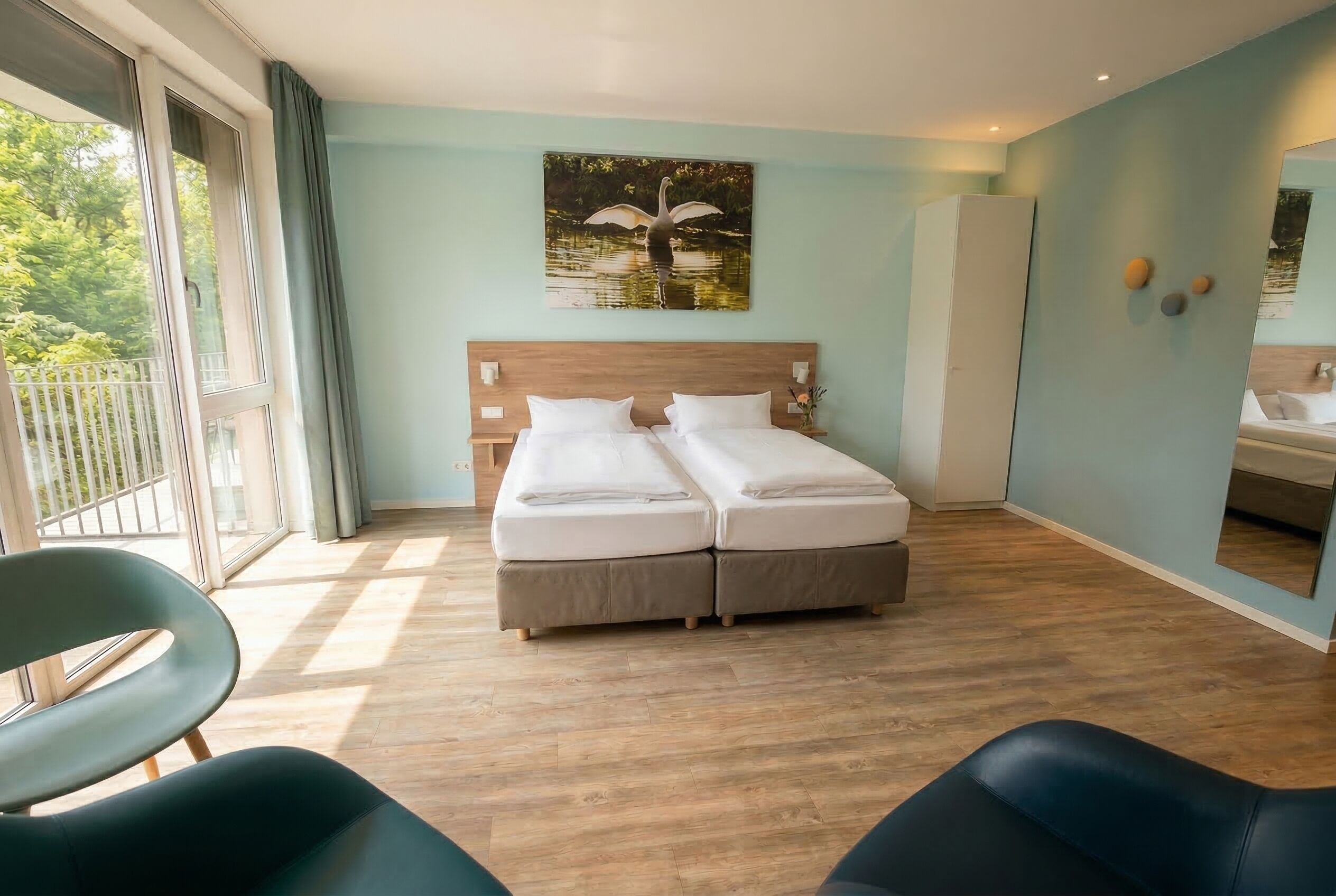 A double bed with white linens, a wooden headboard, a painting of a swan, a balcony with a view of trees, and a bathroom with a mirror and sink.