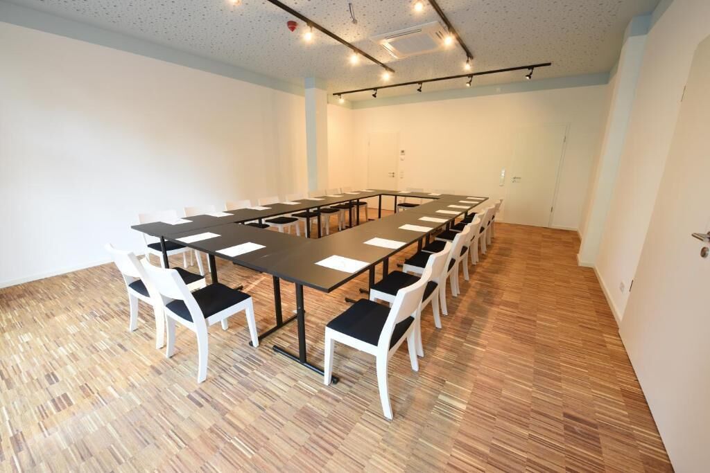 Un salón de conferencias con una larga mesa y sillas blancas, suelo de madera y paredes blancas.