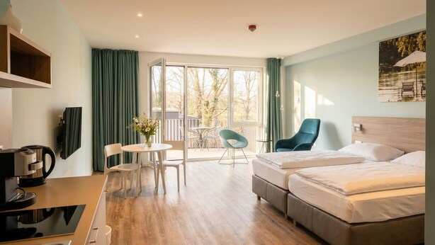 A hotel room with a bed, a desk with a coffee maker, a chair, a small table with flowers, and a balcony with chairs. - Steverbett Hotel (Luedinghausen)