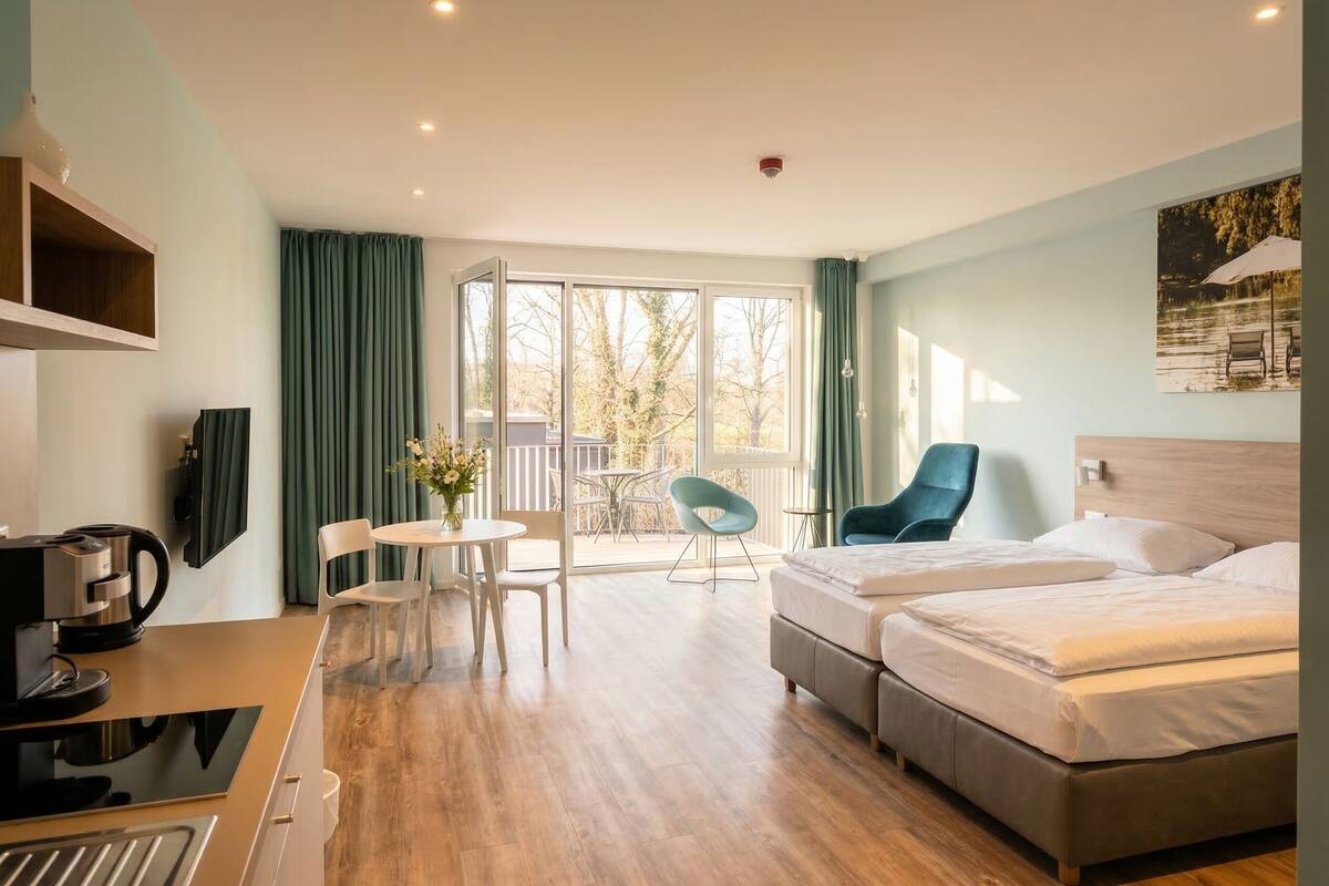 A hotel room with a bed, a desk with a coffee maker, a chair, a small table with flowers, and a balcony with chairs.