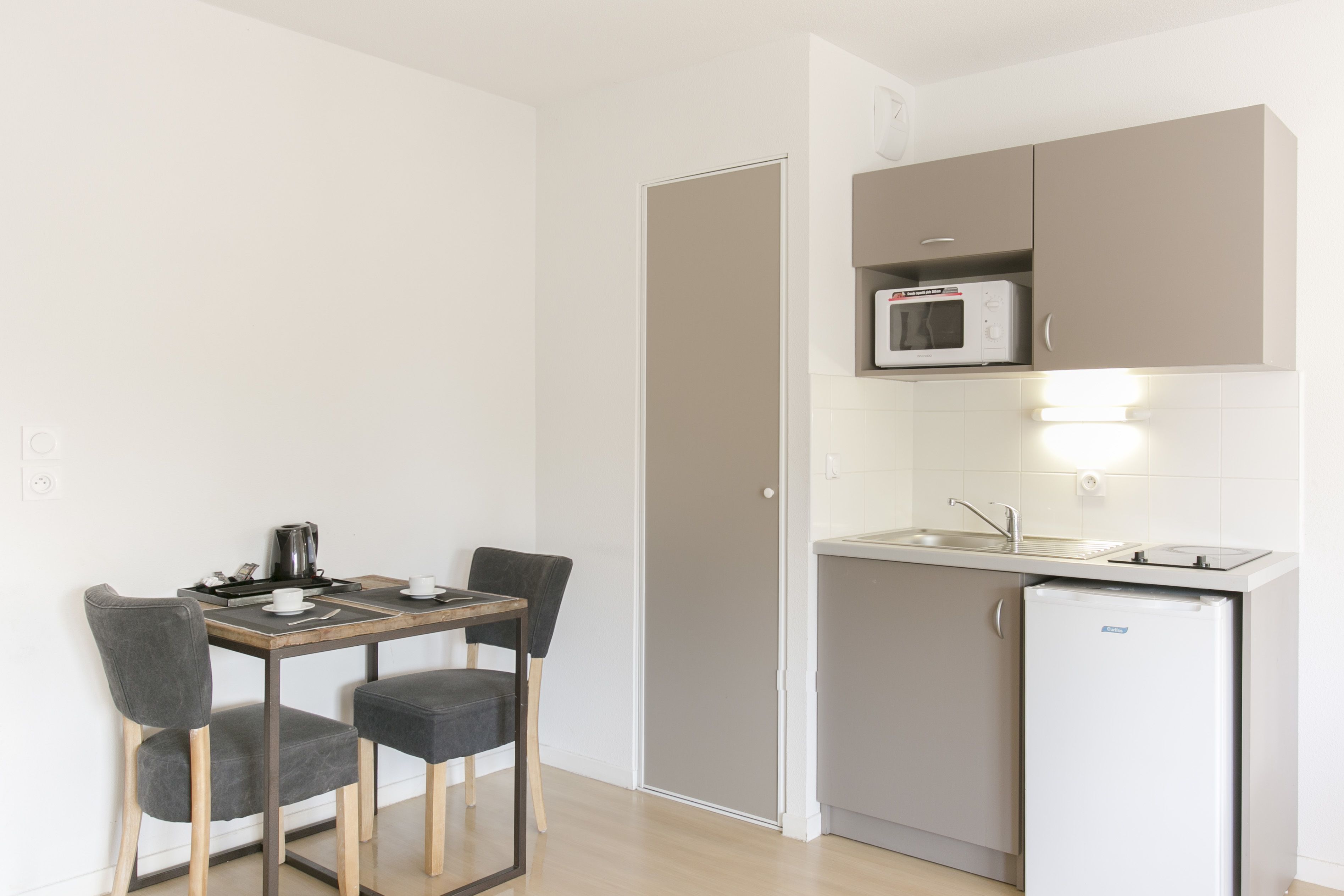 Een kleine keuken met een magnetron, spoelbak en een kleine koelkast. Er staat een eettafel met twee stoelen.