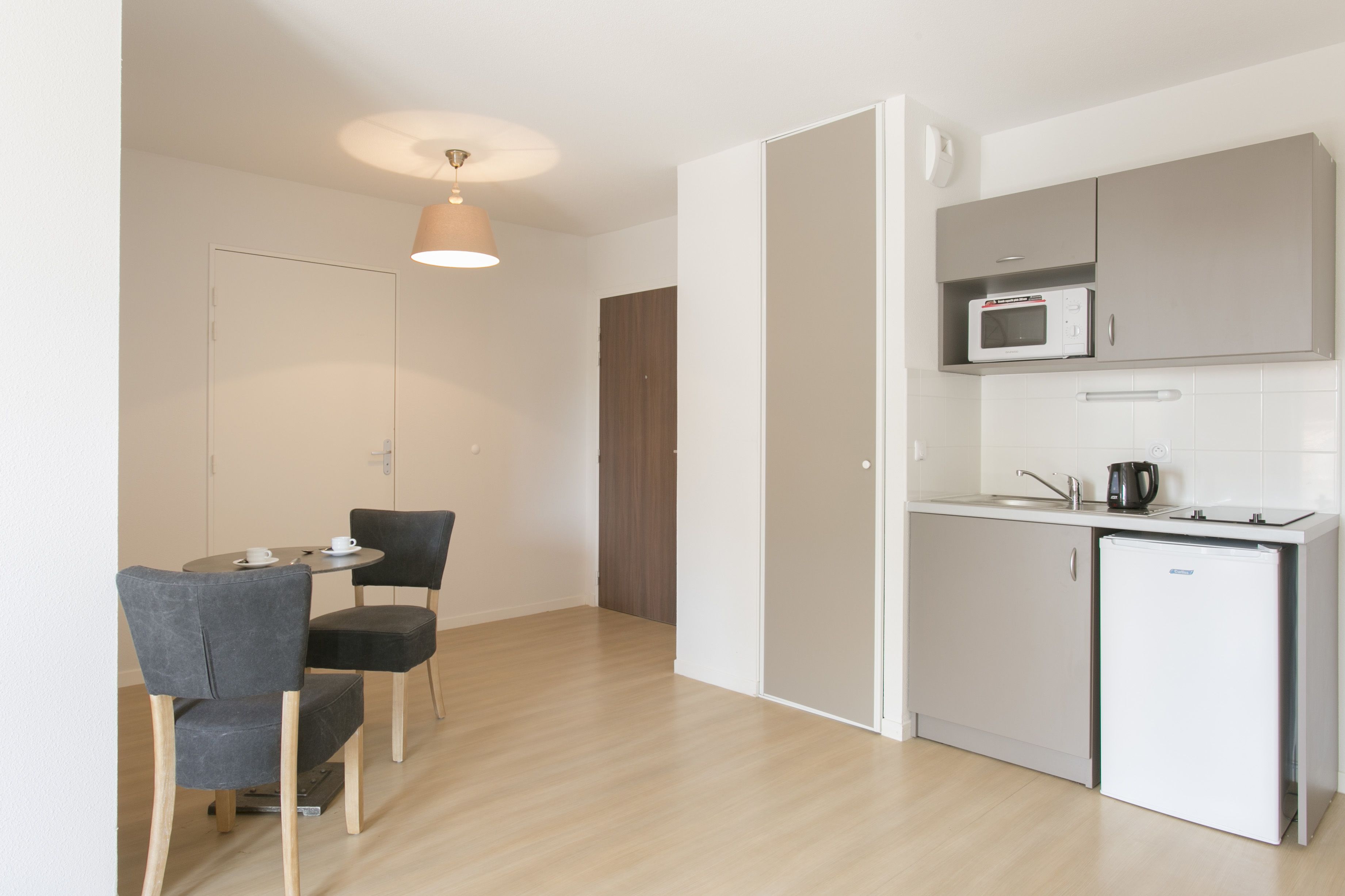 Cocina compacta con microondas, cafetera y refrigerador pequeño. Cuenta con un comedor con dos sillas y una mesa.