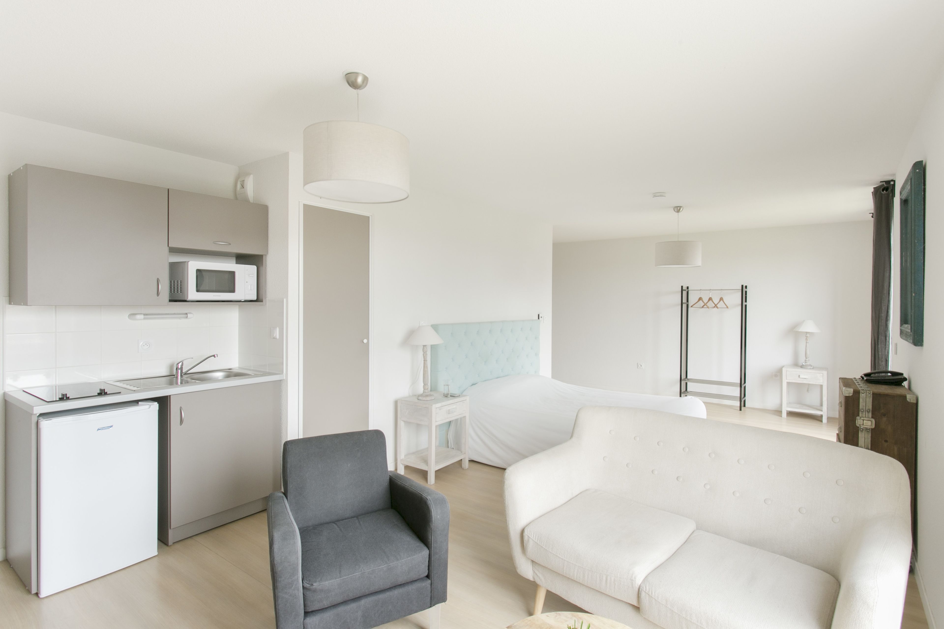 A compact living space with a kitchenette, a gray armchair, a white sofa, and a bed with a headboard.