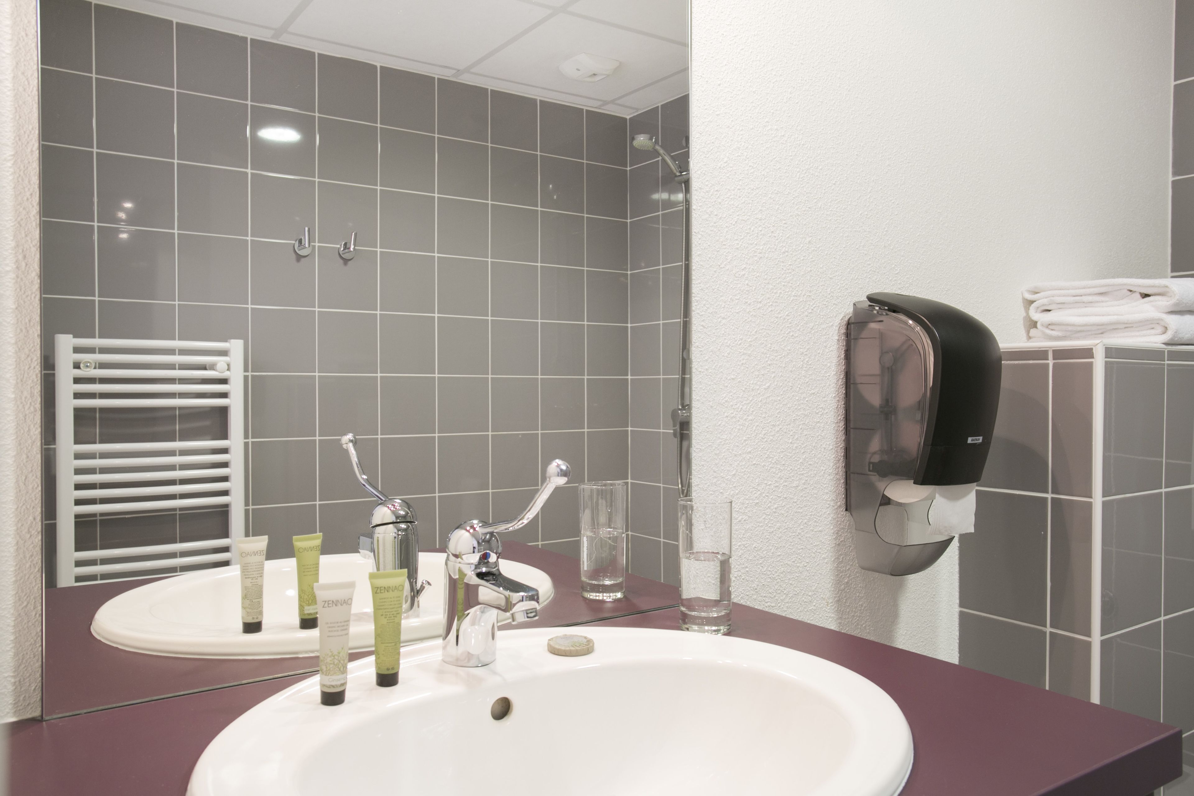 A bathroom with a white sink, two faucets, a mirror, a towel rack, and a soap dispenser.