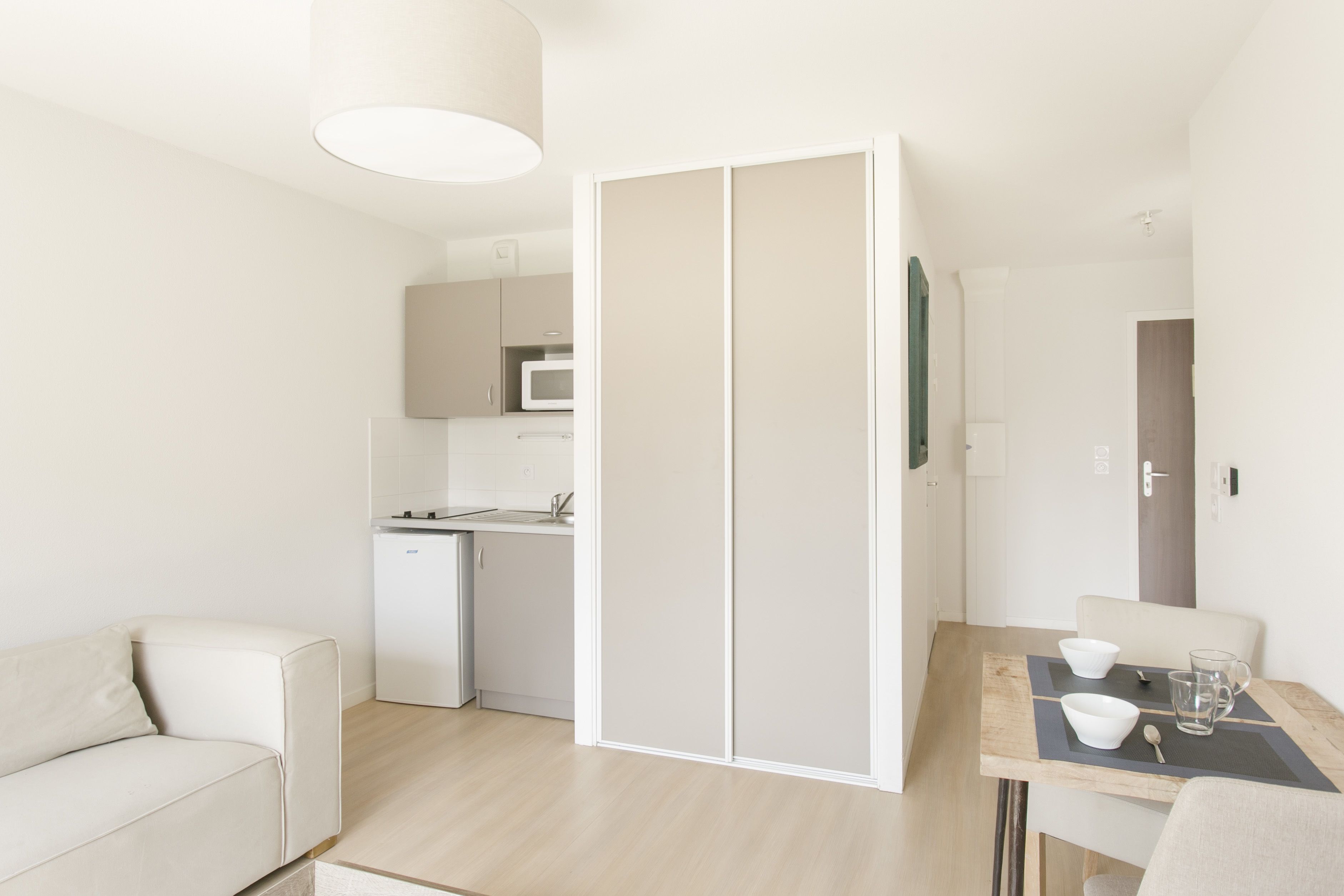 Un espace de vie moderne et minimaliste comprenant un canapé, une table à manger avec quelques éléments de décoration, et un coin cuisine avec des placards et un four à micro-ondes.