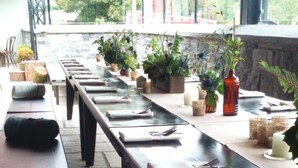 A long dining table set with benches, surrounded by potted plants and candles, in a stone-walled room with a view of trees. - Ryan's Hotel (Cong)