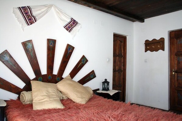 A room with a bed, a wooden headboard with peacock feather designs, a white wall, and a wooden door.