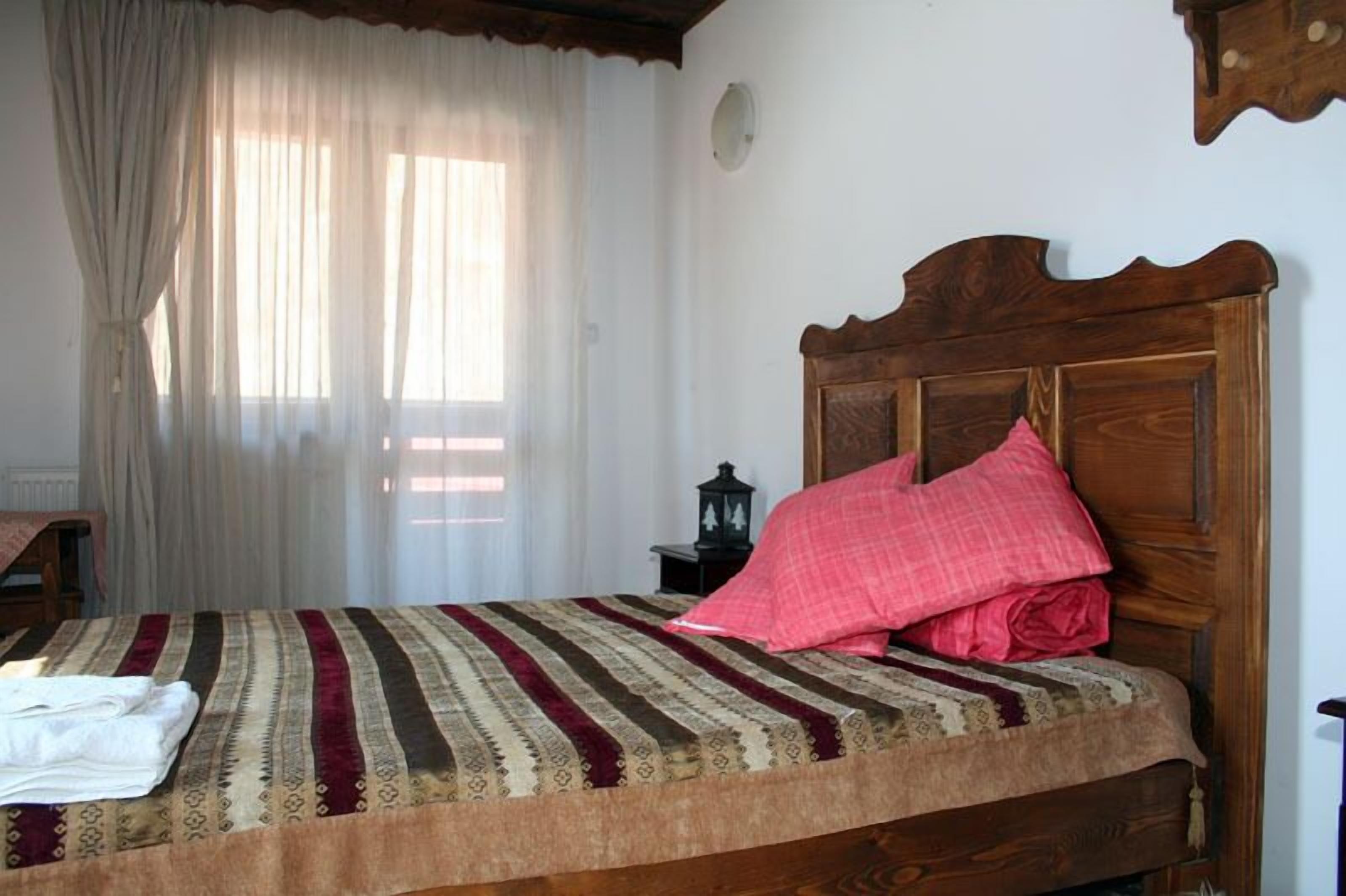 Uma cama com colcha estampada e cabeceira de madeira, um criado-mudo com um candeeiro e uma janela com cortinas leves.