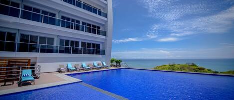 A modern hotel with a large infinity pool, lounge chairs, and a view of the ocean.