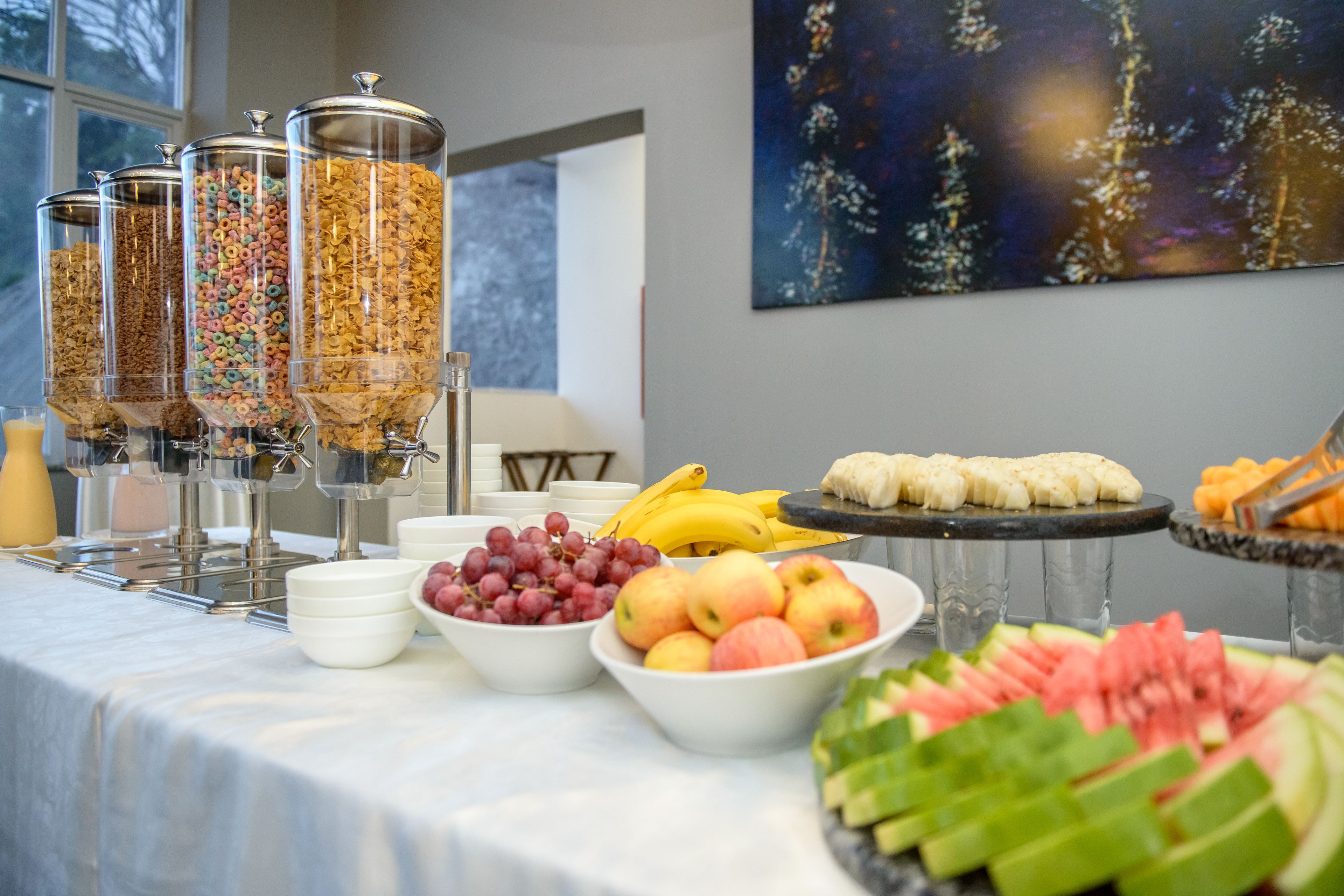 A buffet table with cereal dispensers, bowls of grapes, apples, bananas, and watermelon slices.