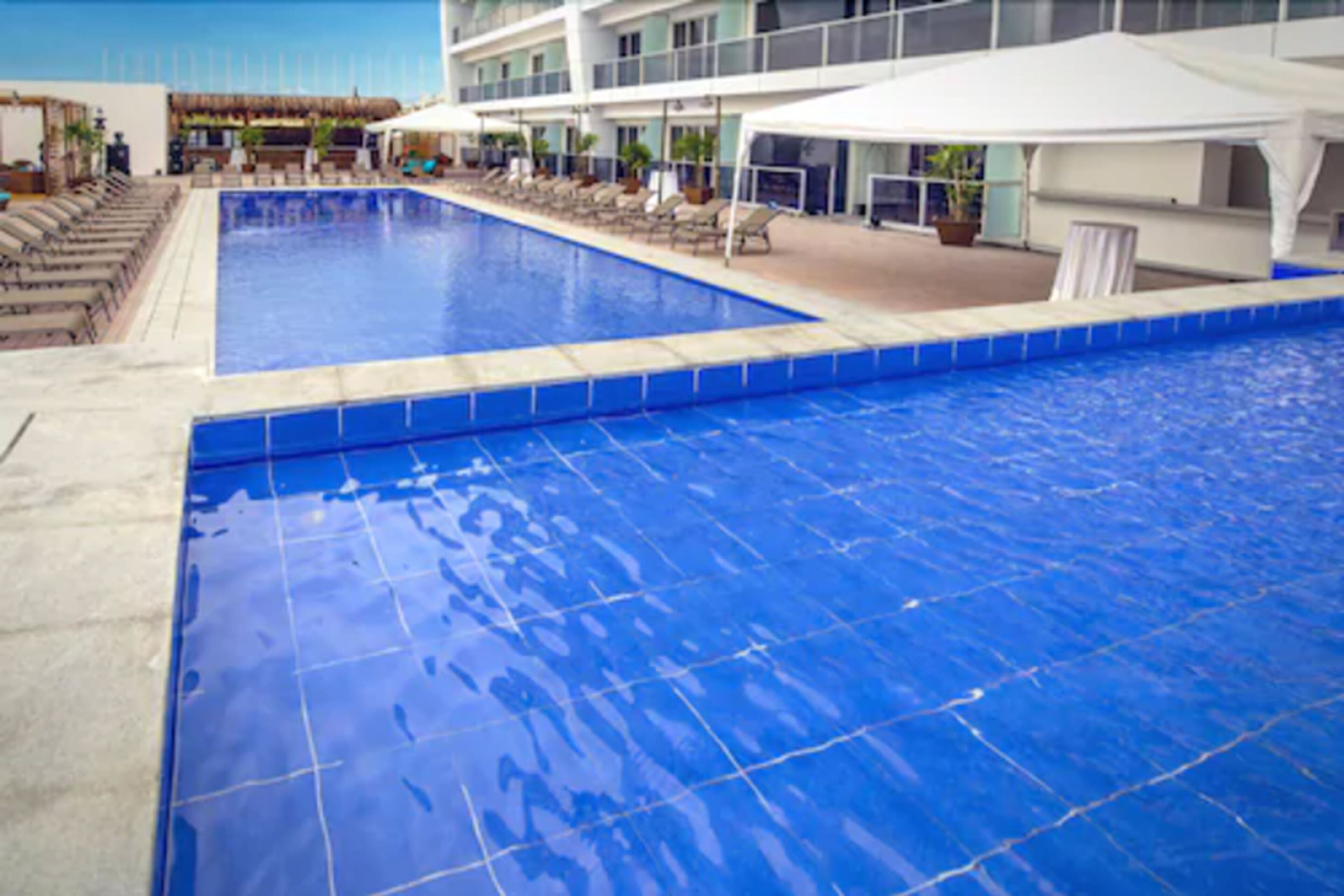 Ein großer, überdachter Pool mit Liegestühlen und Sonnenschirmen, umgeben von einem Gebäude mit mehreren Etagen und großen Fenstern.