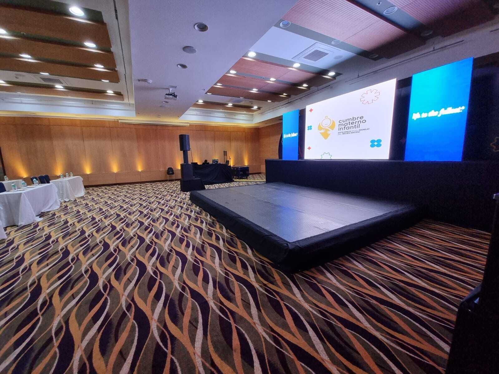 Une salle de conférence dotée d’une scène, de rangées de tables avec des chaises et d’un tapis à motifs.