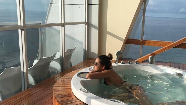 Una persona relajándose en un jacuzzi sobre una terraza de madera con amplios ventanales que ofrecen vista al mar.